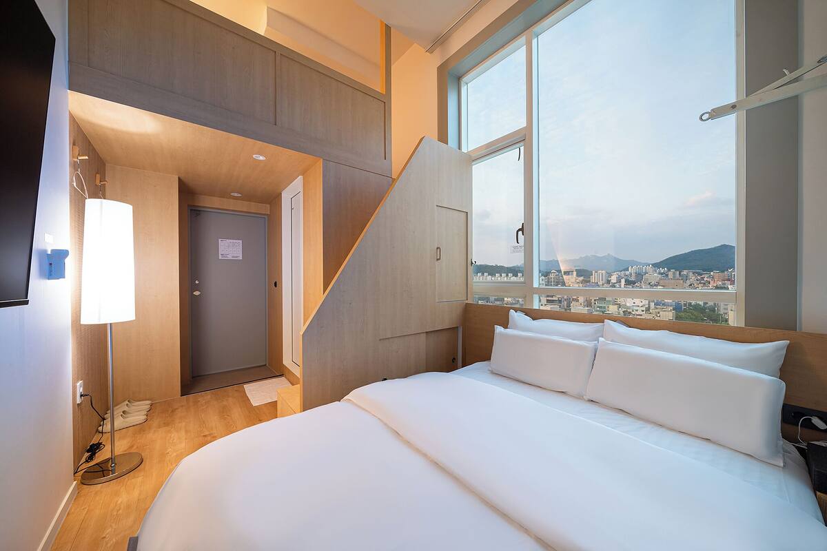 A hotel room with a bed, a desk, a lamp, a window with a city view, and a door leading to another room.