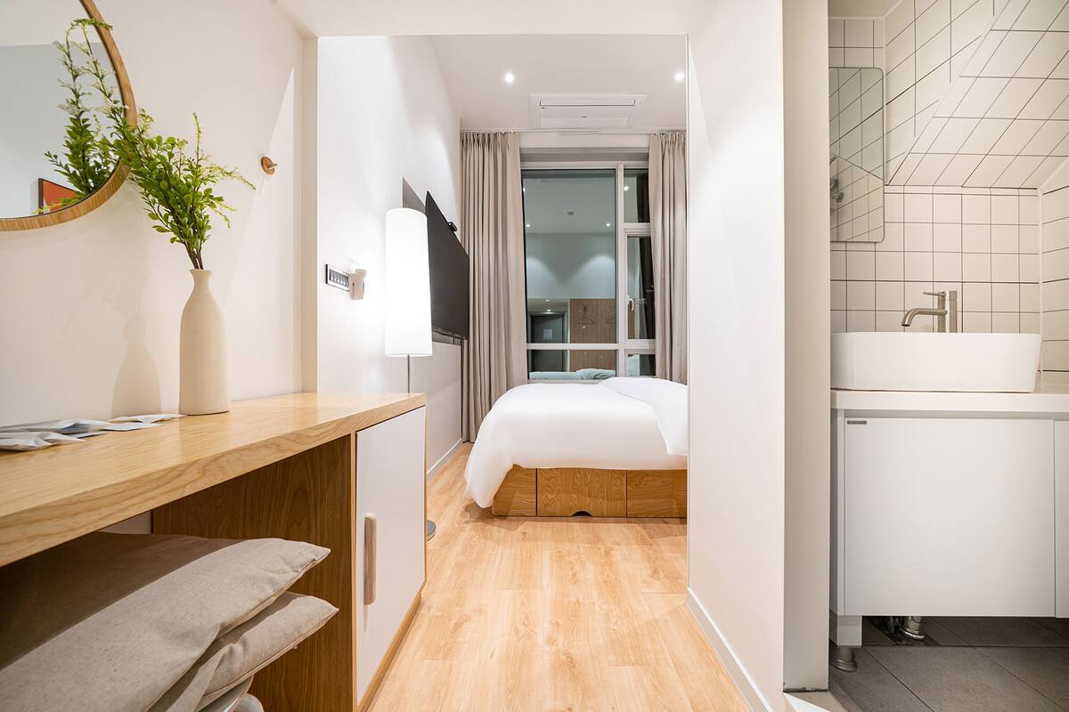 A hotel room with a wooden desk, a bed with a white mattress, a mirror, a plant, and a bathroom with a sink and tiled walls.
