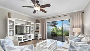 Una sala de estar moderna con un televisor de pantalla plana, muebles blancos, un ventilador de techo y un ventanal corredizo al exterior.