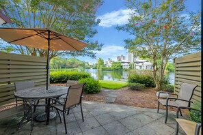 Eine Terrasse mit Tisch und Stühlen, einem Sonnenschirm, Blick auf einen See und einem Gebäude im Hintergrund.
