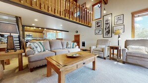 A spacious living room with a high wooden ceiling, a sofa set, a coffee table, and a view of the kitchen in the background.