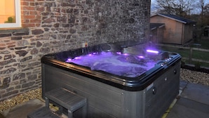A hot tub with a stone wall backdrop, a wooden bench, and a clear view of the surrounding yard and house.