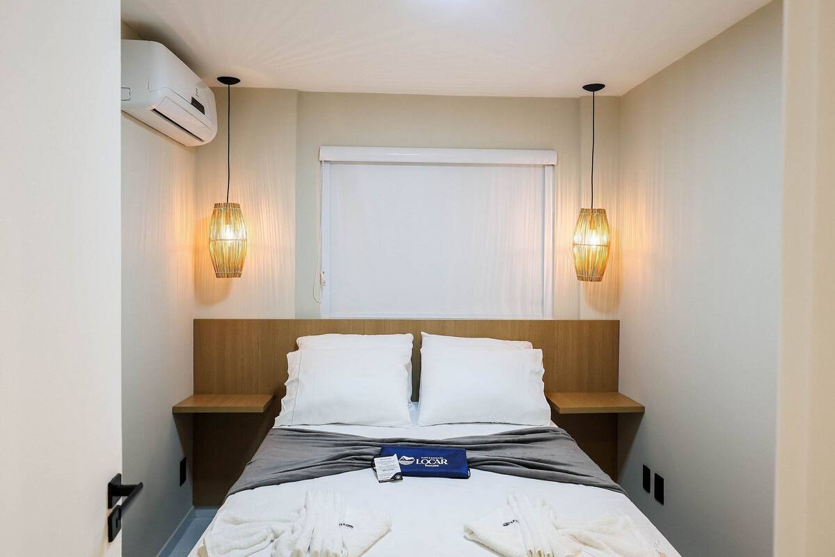 A hotel room with a bed, bedside tables, a nightstand, an air conditioning unit, and two wall-mounted lamps.