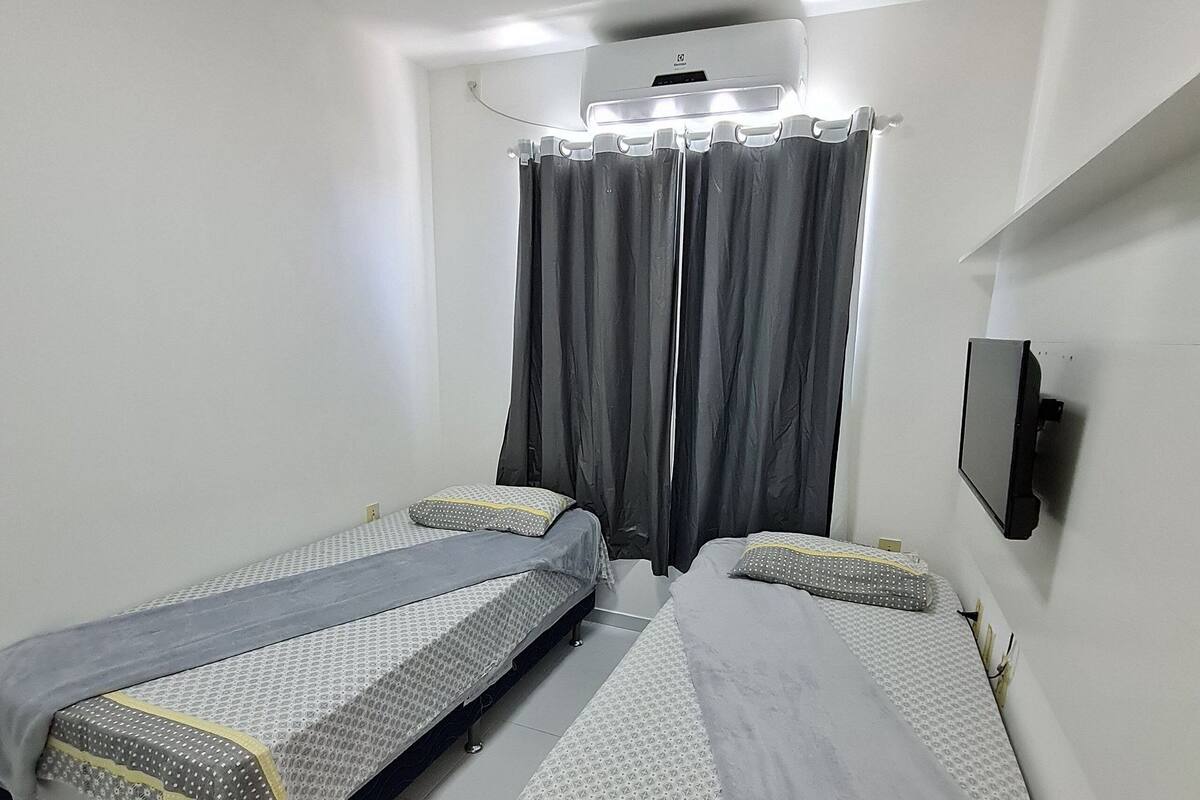 A small, clean room with two single beds, a TV mounted on the wall, and an air conditioning unit above the beds.