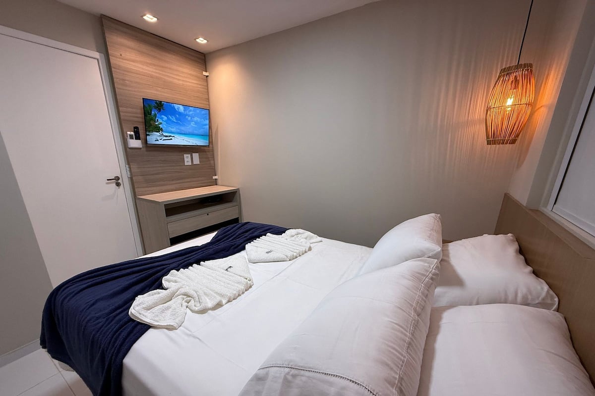 A neatly made bed with white linens and a blue throw blanket, a wooden nightstand with a flat-screen TV, and a wall-mounted light fixture.
