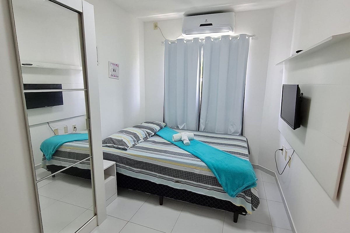 A small, clean room with a bed, a mirror, a TV, and a window with curtains.