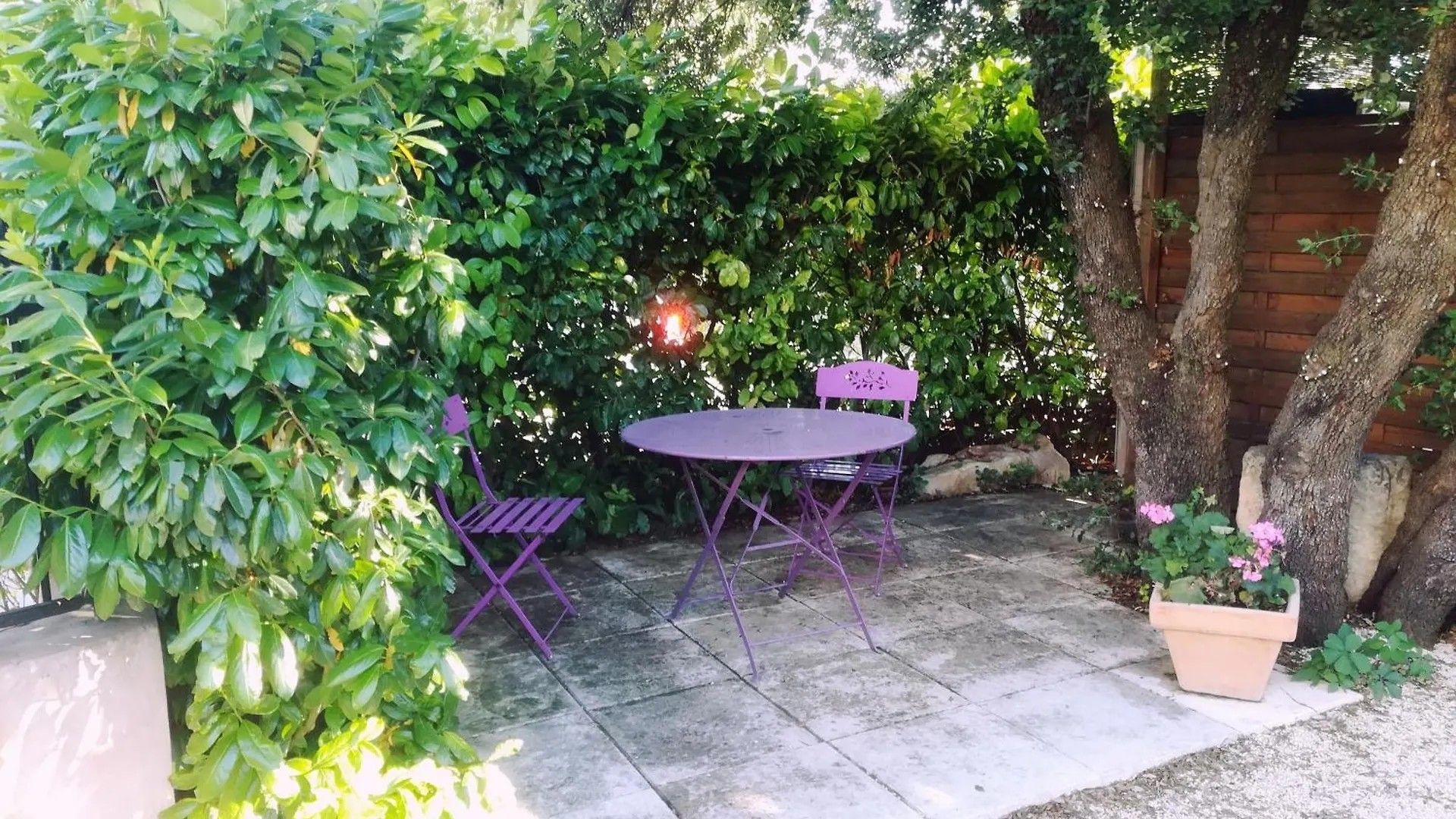 Une terrasse extérieure avec une table ronde et deux chaises, entourée d’une végétation luxuriante et d’une clôture en bois.