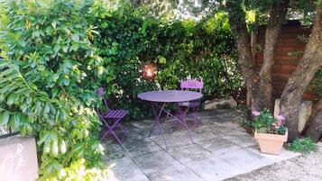 Une terrasse extérieure avec une table ronde et deux chaises, entourée d’une végétation luxuriante et d’une clôture en bois.