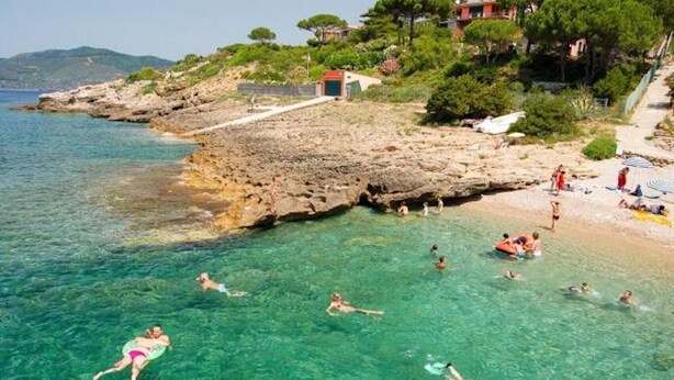 Una playa costera con agua cristalina, costa rocosa y gente nadando.