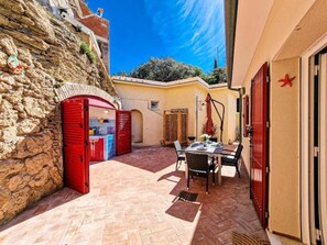 Un muro in pietra, una porta rossa, un tavolo con sedie e una piccola finestra.