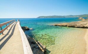 Um cais que se estende sobre águas azul-claras, com uma costa rochosa e colinas ao longe.