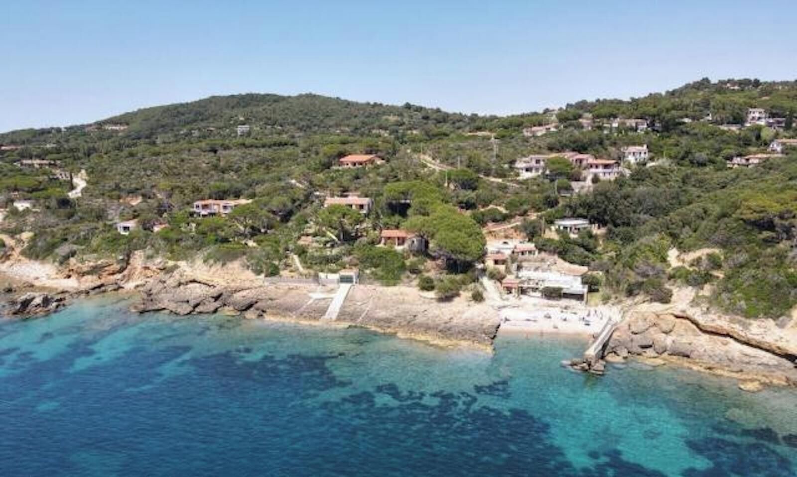 A coastal area with buildings on a hillside, a rocky shoreline, and clear blue water.