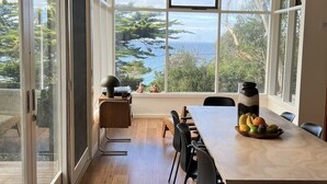 Un comedor con mesa y sillas de madera, un ventanal con vista al mar y una pequeña mesa con frutas y una botella de agua.
