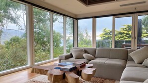 Un salón moderno con un sofá grande, una mesa de centro de madera y vistas a zonas verdes a través de amplios ventanales.