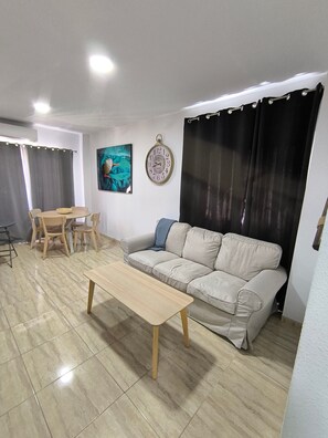 A living room with a sofa, a coffee table, a dining table with chairs, a wall clock, and a painting.