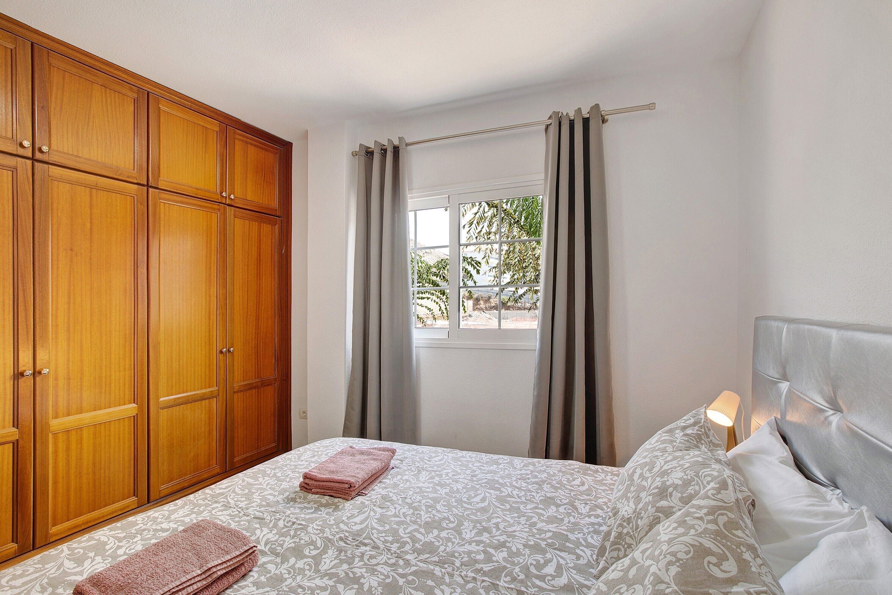 Ein Schlafzimmer mit Bett, Kleiderschrank, einem Fenster mit Vorhängen und einem Nachttisch mit Lampe.