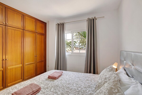 A bedroom with a bed, a wardrobe, a window with curtains, and a nightstand with a lamp.