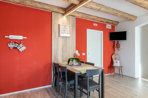 Un coin repas avec un mur rouge, des poutres en bois au plafond, un téléviseur fixé au mur et une table avec des chaises.