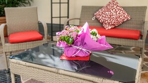 Ein Korbstuhl mit rotem Kissen, ein Glastisch mit Blumenstrauß und ein gemustertes Kissen auf einer Couch.