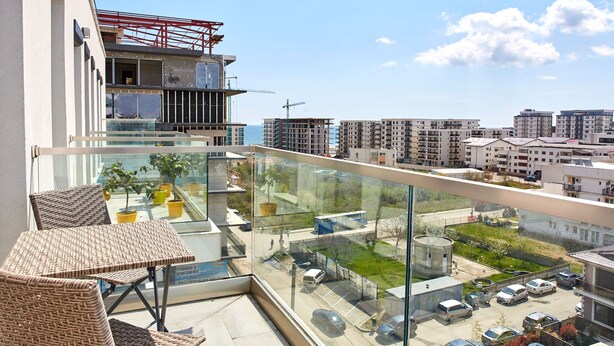 Varanda com mobiliário de vime, guarda-corpo de vidro e vista para um parque de estacionamento e edifícios.