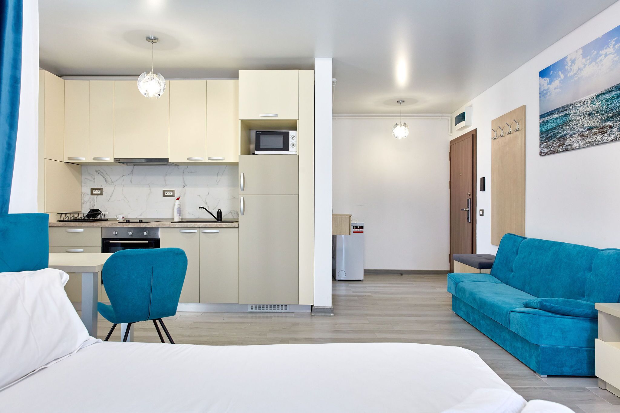 Een moderne hotelkamer met een bed, een blauwe fauteuil, een blauwgroene bank, een kitchenette en een schilderij van de zee aan de muur.