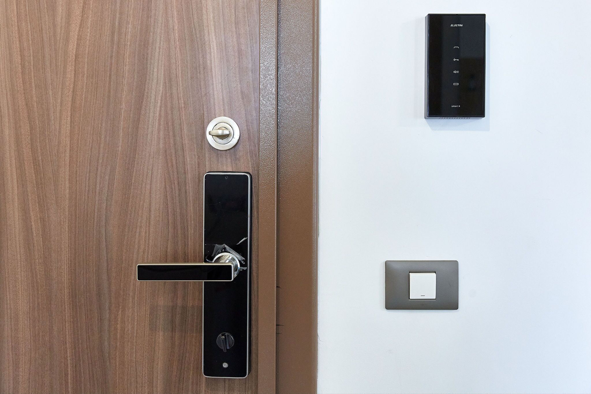 Een houten deur met een modern slot en een bedieningspaneel aan de muur.