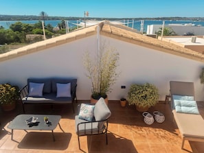A rooftop terrace with a sofa, a small table, potted plants, and a view of the sea.