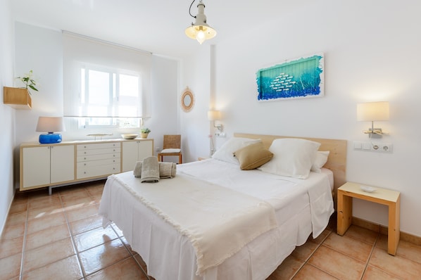 A bedroom with a large bed, a bedside table, a nightstand, a dresser, a mirror, a wall art piece, and a window.