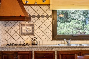 A kitchen with a gas stove, a kettle, a sink, and a window with a view of greenery.