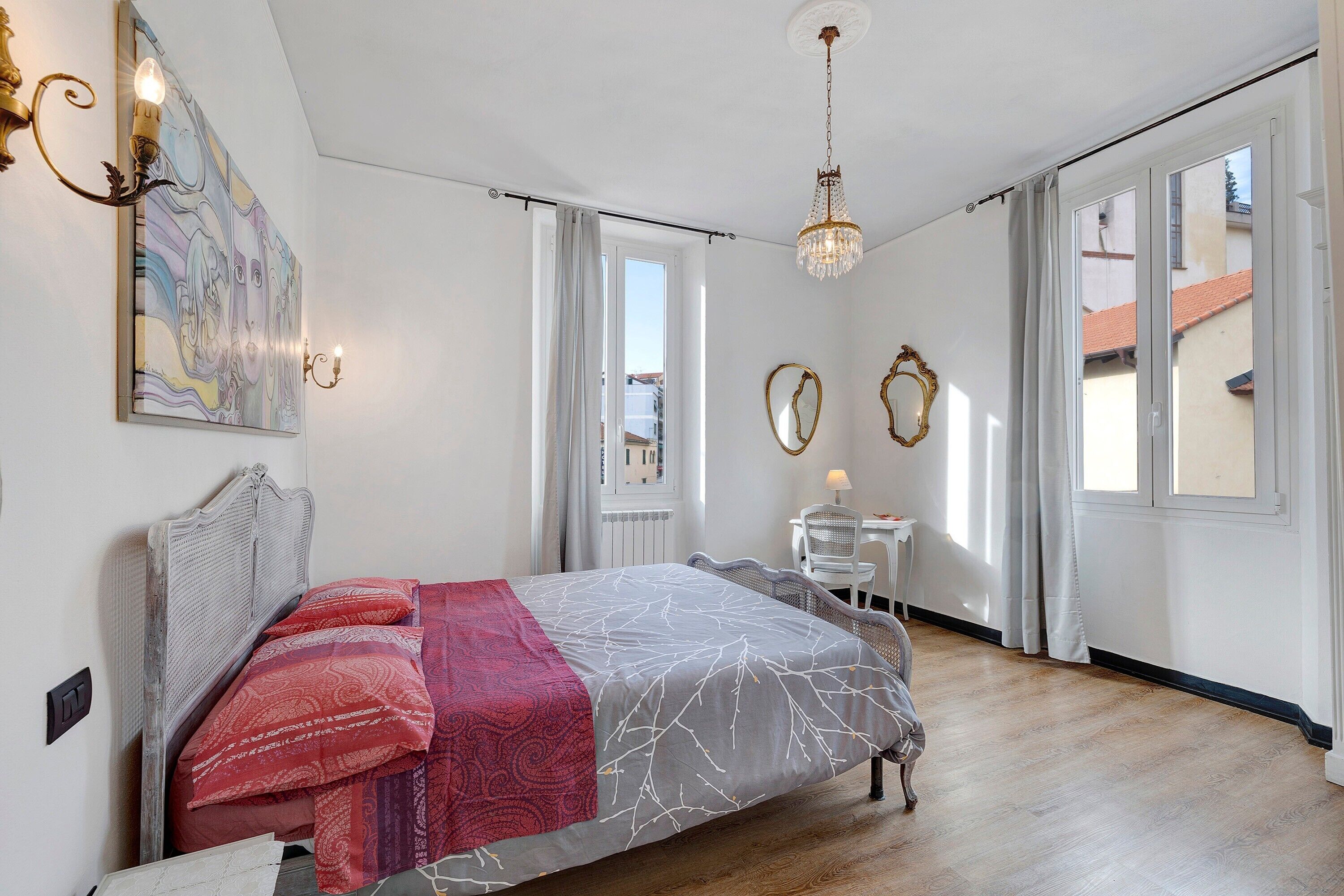 Quarto com cama, escrivaninha, cadeira, lustre e vista para edifícios pela janela.