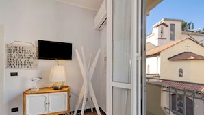 Quarto com televisão, suporte branco, armário de madeira e varanda com guarda-corrimão.