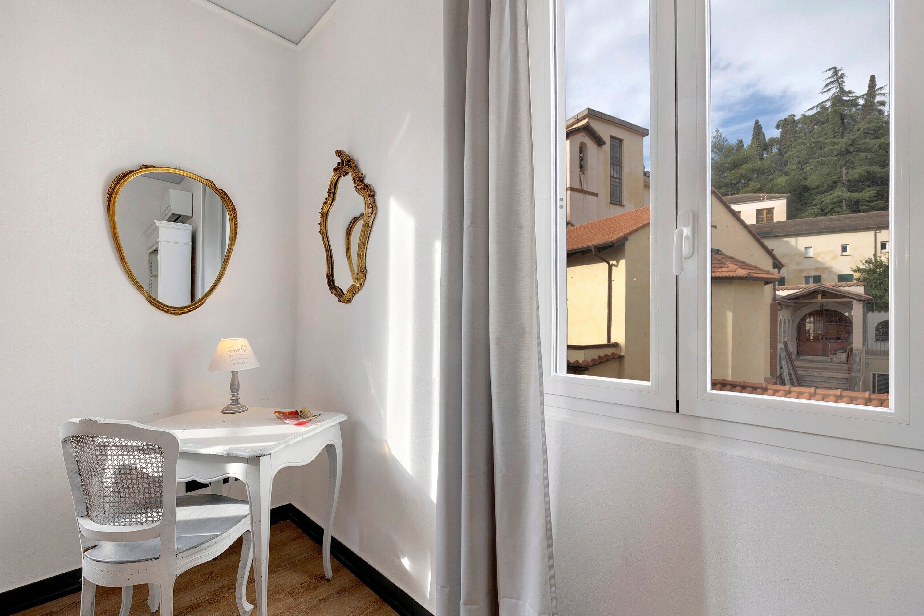 A room with a white table, a chair, a lamp, two wall mirrors, and a window with a view of buildings and trees.