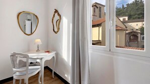 Quarto com uma mesa branca, uma cadeira, um abajur, dois espelhos na parede e uma janela com vista para edifícios e árvores.