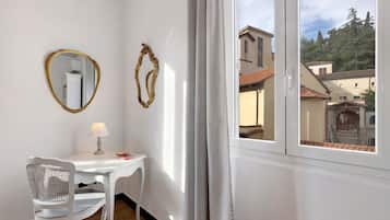 A room with a white table, a chair, a lamp, two wall mirrors, and a window with a view of buildings and trees.