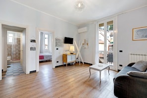 Un salon moderne avec un plancher en bois, un canapé, une table basse et un téléviseur. On aperçoit une porte donnant accès à une salle de bain et un balcon à travers une porte ouverte.