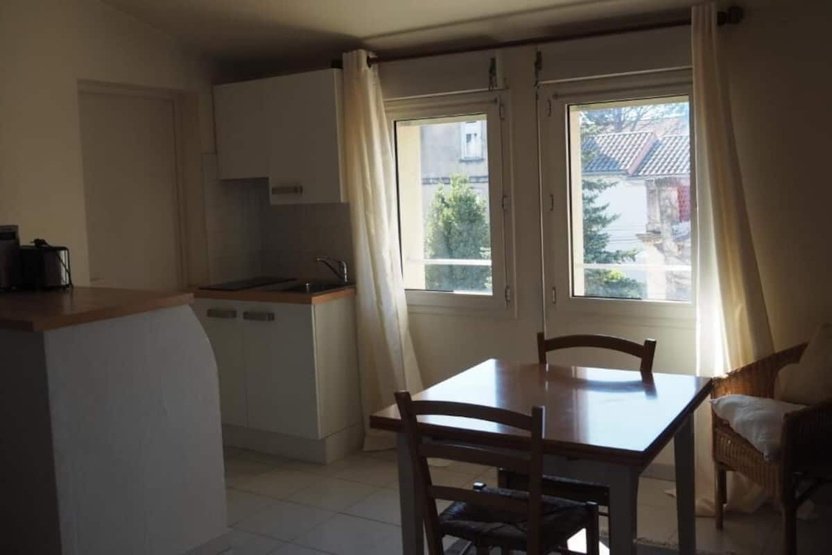 A compact kitchen with a dining area, featuring a large window with curtains, a built-in oven, and a small countertop.