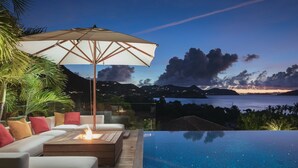 Une piscine à débordement agrémentée d’un foyer extérieur, de coins salon en plein air et d’un grand parasol offrant de l’ombre.
