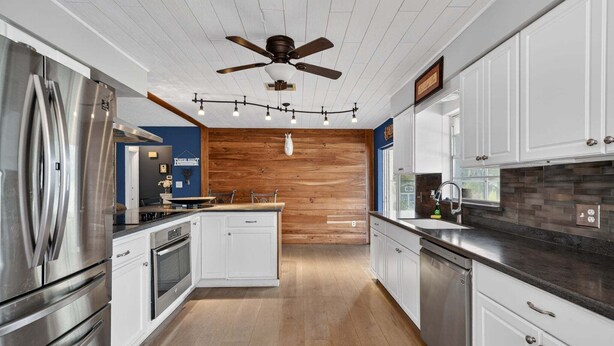 A modern kitchen with stainless steel appliances, white cabinetry, a wooden accent wall, and a ceiling fan. - The Lake Home - Beautiful Oasis In The Heart Of Florida! 2 Bedroom Home by RedAwning (Haines City)