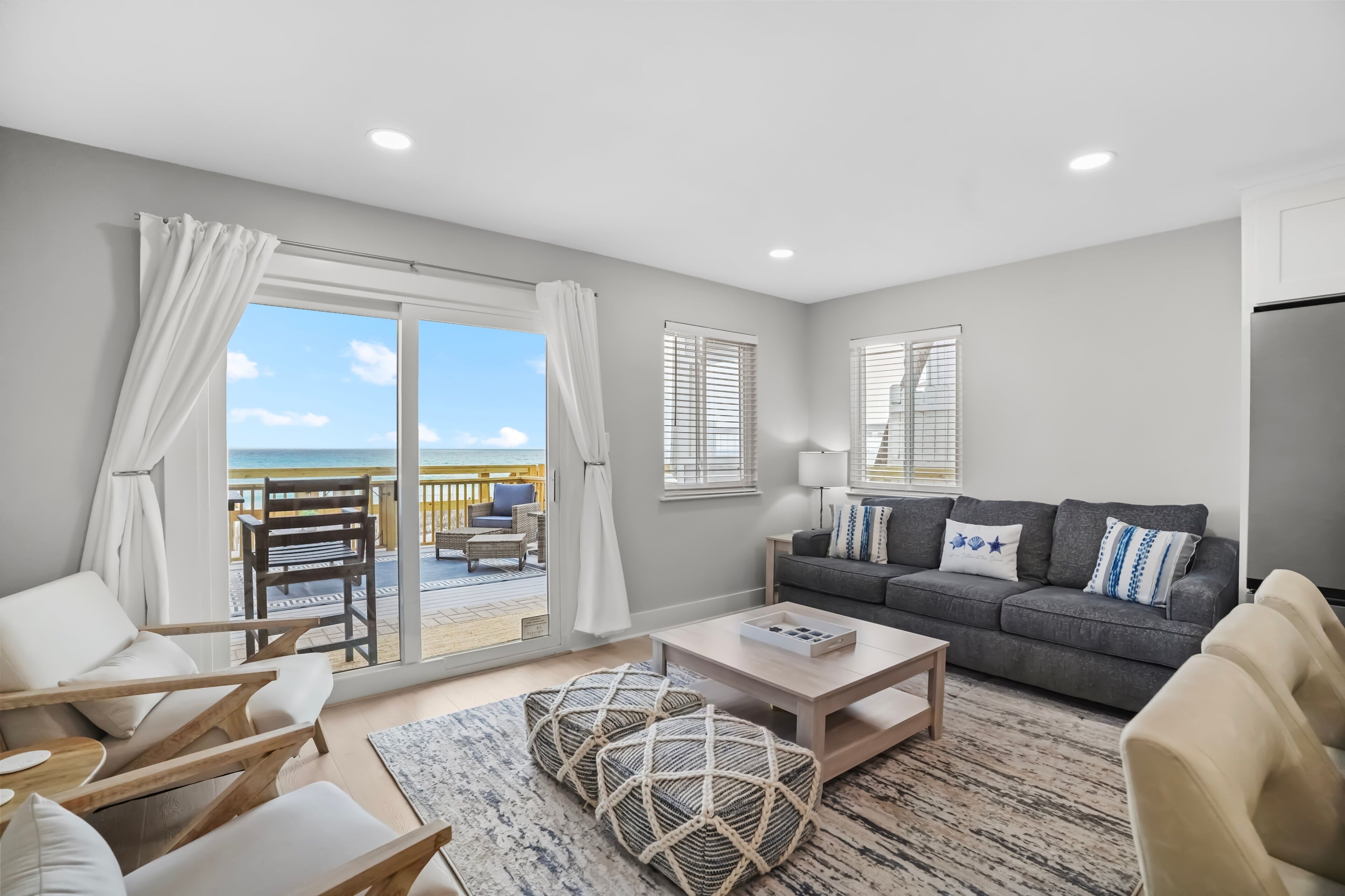 A modern living room with a sofa, coffee table, and chairs, a balcony with a view of the sea, and a large window with blinds.
