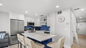 A modern kitchen with white cabinets, a stainless steel refrigerator, a blue tiled backsplash, and a central island with bar stools. - Leeward Ii-3-front Row 1 Bedroom Condo by RedAwning (Santa Rosa Beach)