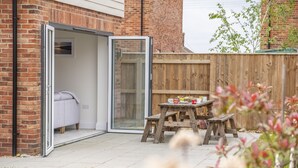 A patio with a sliding glass door leading to a room with a bed and a white chair. There is a wooden table with cups and a small stool.