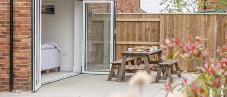 A patio with a sliding glass door leading to a room with a bed and a white chair. There is a wooden table with cups and a small stool.
