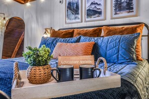 A bed with a sign reading 'Adventure Awaits,' a potted plant, and two black mugs on a tray. - Cliff Haven 1 Bedroom Cabin by RedAwning (Townsend)