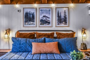 Una cama con colcha azul y almohadillas naranjas, una obra de arte enmarcada encima y lámparas fijadas en la pared a cada lado.