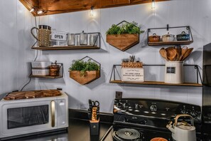 A kitchen with white cabinets, a microwave, a stove, and various utensils and containers on shelves. - Cliff Haven 1 Bedroom Cabin by RedAwning (Townsend)