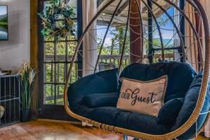 A hanging chair with a cushion labeled 'OUR GUEST' in a room with large windows and a view of greenery outside. - Cliff Haven 1 Bedroom Cabin by RedAwning (Townsend)