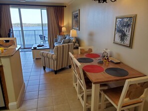A cozy living area with a striped armchair, a sofa, and a dining table with placemats. There is a large window with curtains, a lamp, and framed pictures on the wall.