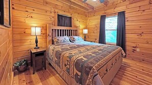 A wooden cabin bedroom with a bed, two bedside lamps, a ceiling fan, and a window with curtains. - High Mountain Cabin Long Range Mountain Views at 4,000 Ft above Sea Level (Bryson City)
