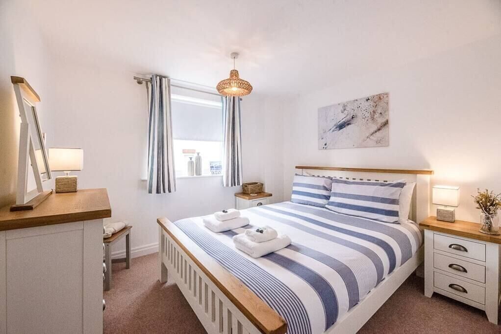 A bedroom with a bed, bedside tables, a window with curtains, a hanging light fixture, and a wall-mounted artwork.