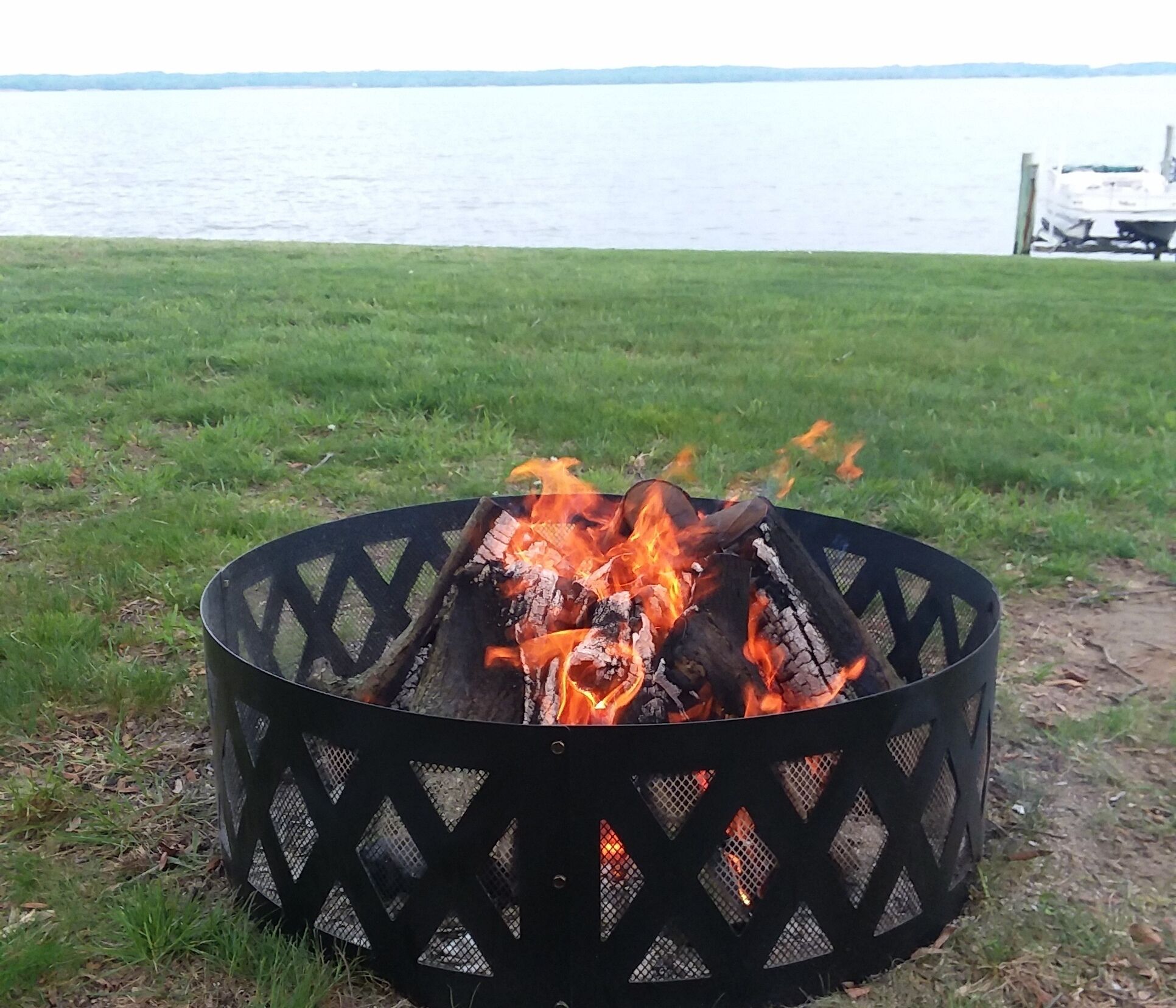 Ein schwarzer Metall-Feuerkorb mit brennenden Holzscheiten, vor einer grasbewachsenen Fläche mit einem Gewässer und einer Anlegestelle.